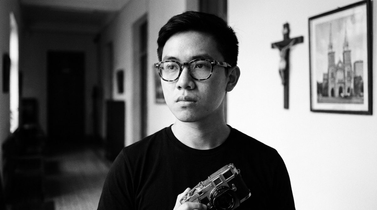 Minh Trần, close-up, holding a Leica — the face behind the voice.